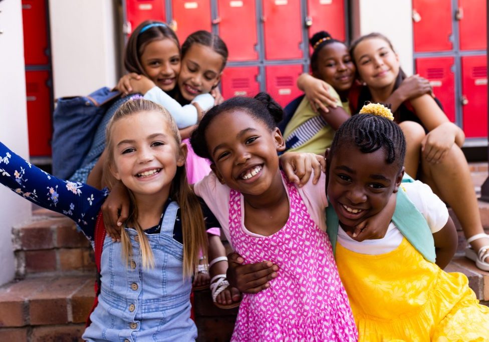 Portrait of diverse happy schoolgirls embracing in elementary school cloakroom. School, learning, childhood, friendship and education, unaltered.