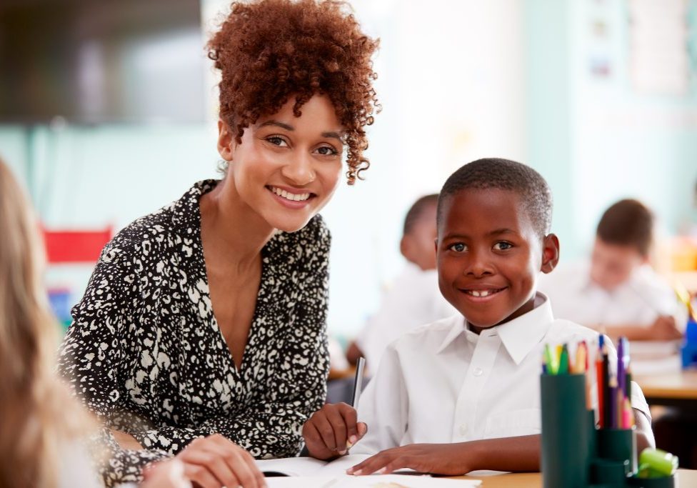 Portrait Of Woman Elementary School Teacher Giving Male Pupil Wearing Uniform One To One Support